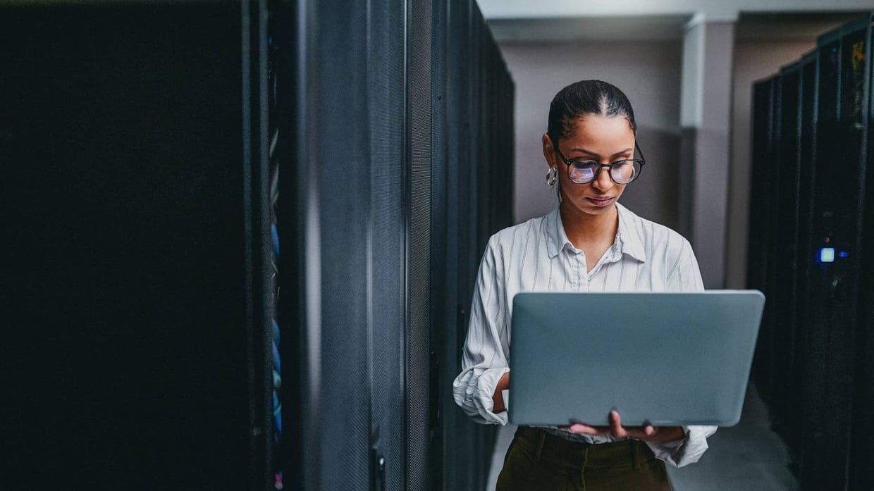 A young woman using a laptop while working in a server room.