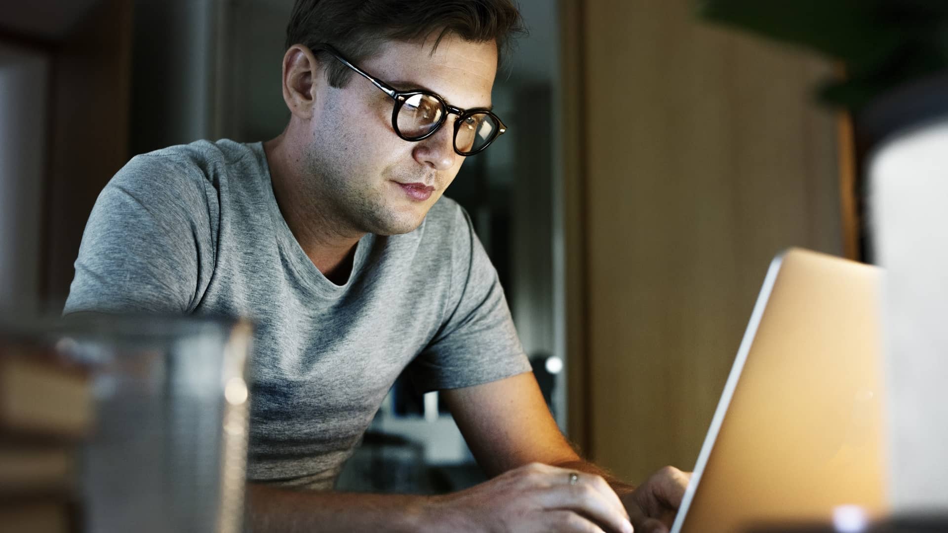 A man wearing glasses and a grey t-shirt working on a laptop at home in the evening.