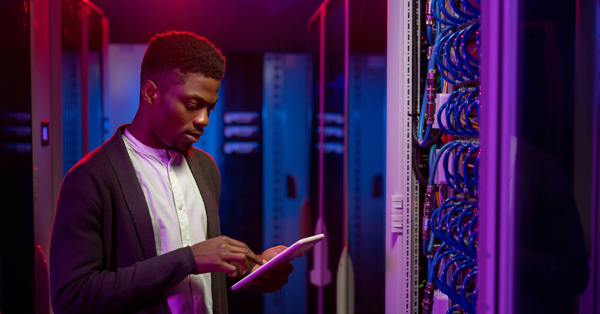 Focused young African-American data engineer standing at open server rack cabinet and using tablet while setting up system at data center.