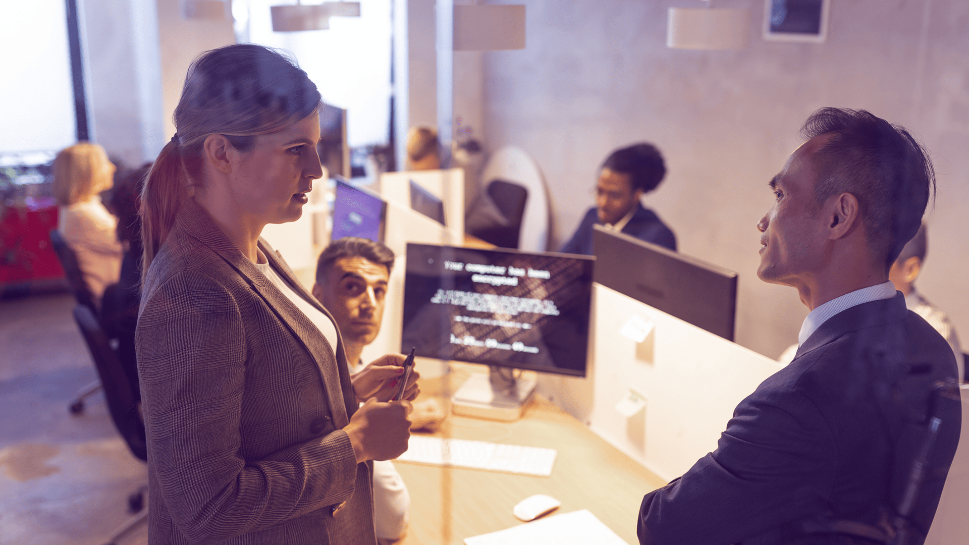 Two corporate office workers discussing ransomware encrypted computers in the occupied glass-walled meeting room behind them.