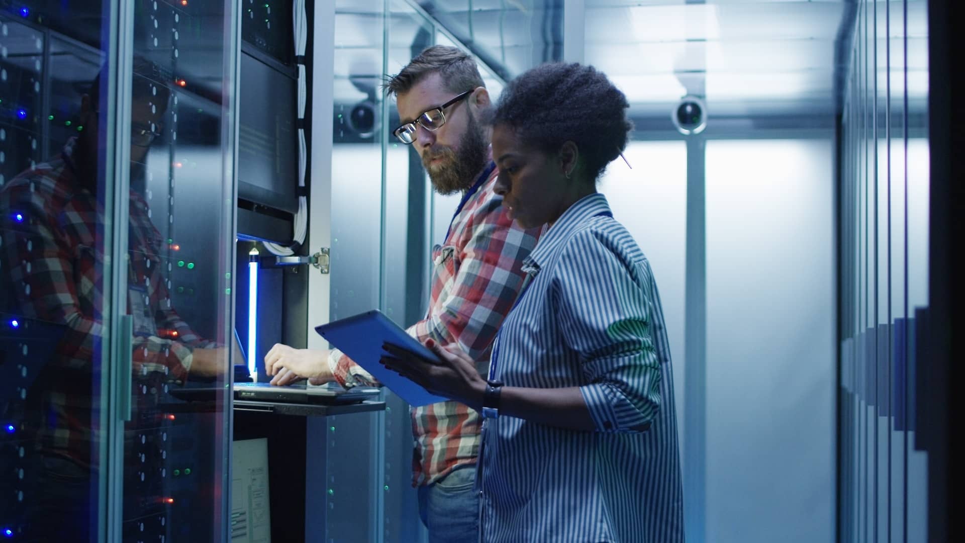 Modern multiethnic man and woman with tablet using laptop in server room while checking servers.