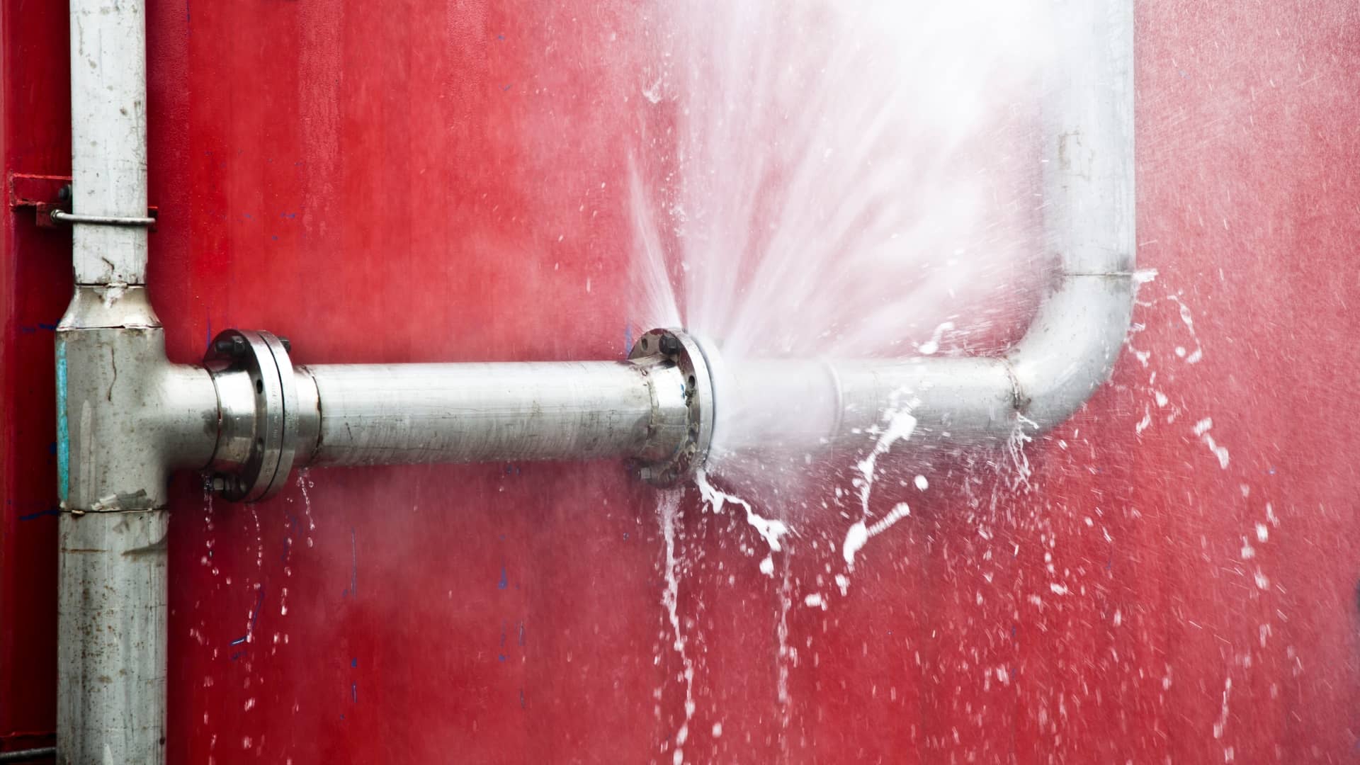 A burst high-pressure metal pipe spraying water against a red wall.